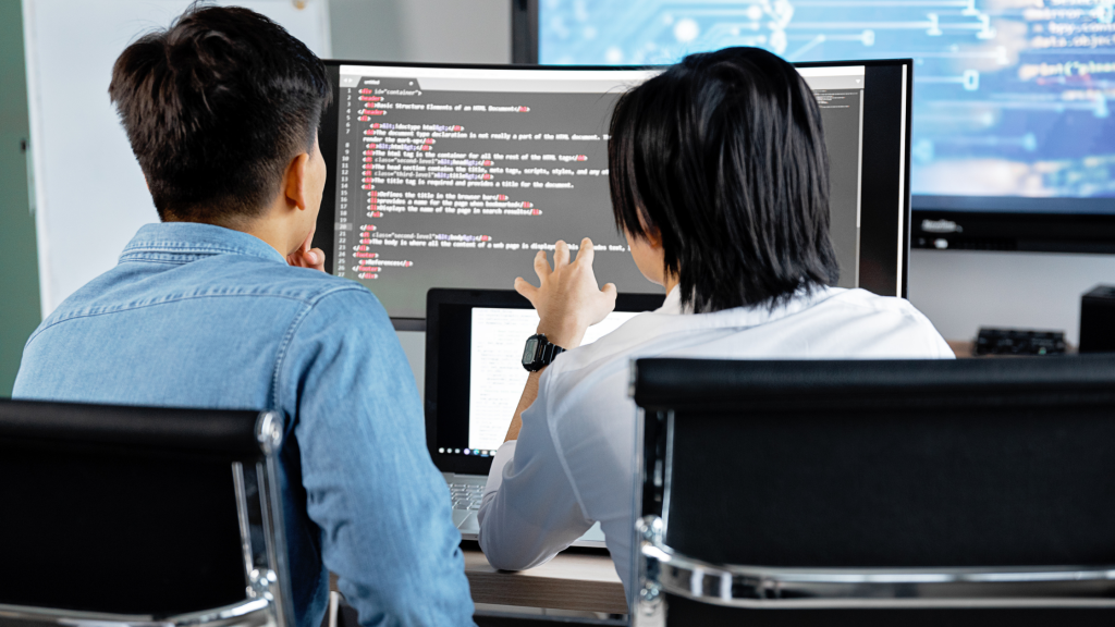 Two programmers collaborate at a desk, viewing code on a large monitor in an office setting.