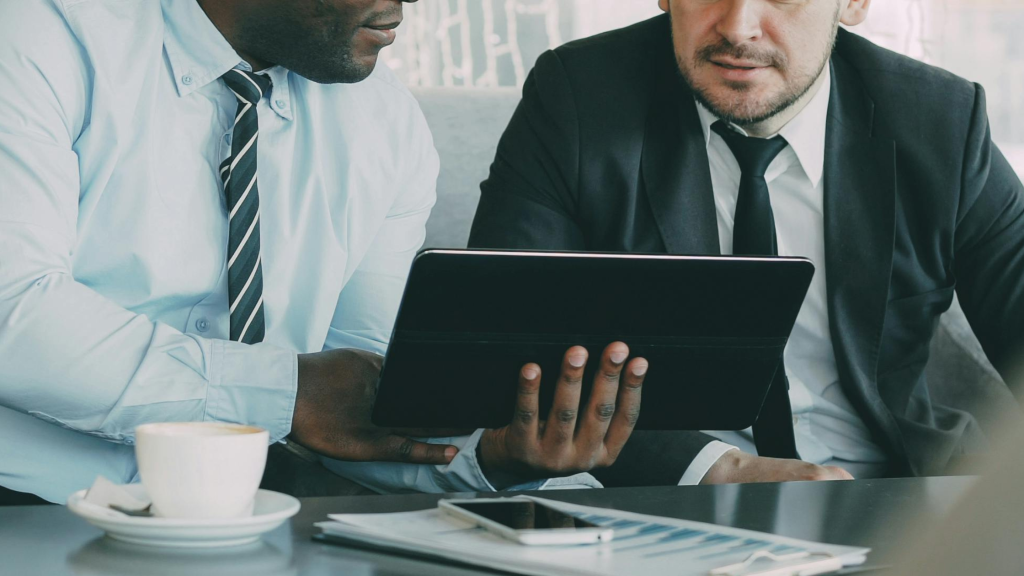 Two businessmen in suits review a tablet together at a table, coffee cup in the foreground.