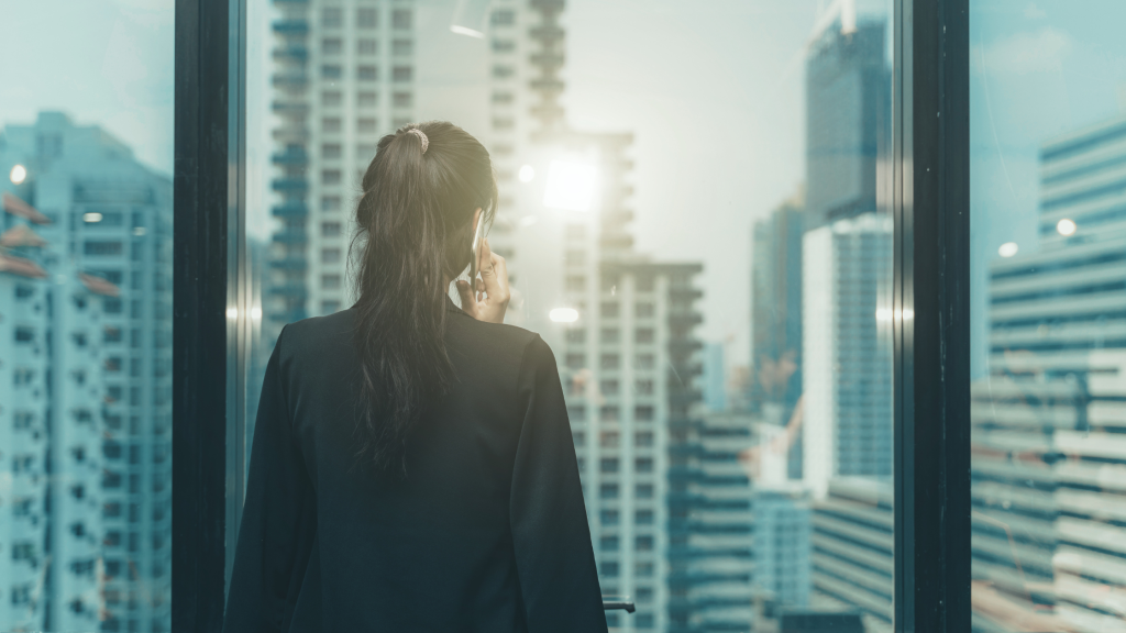 Back view of a person in a dark blazer talking on a mobile phone beside glass walls with a city skyline beyond.