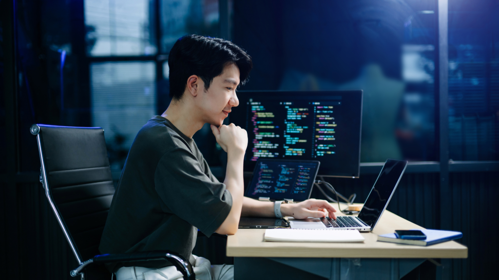 Person coding at a desk in a dim, blue-toned office, with a large monitor displaying colorful code.