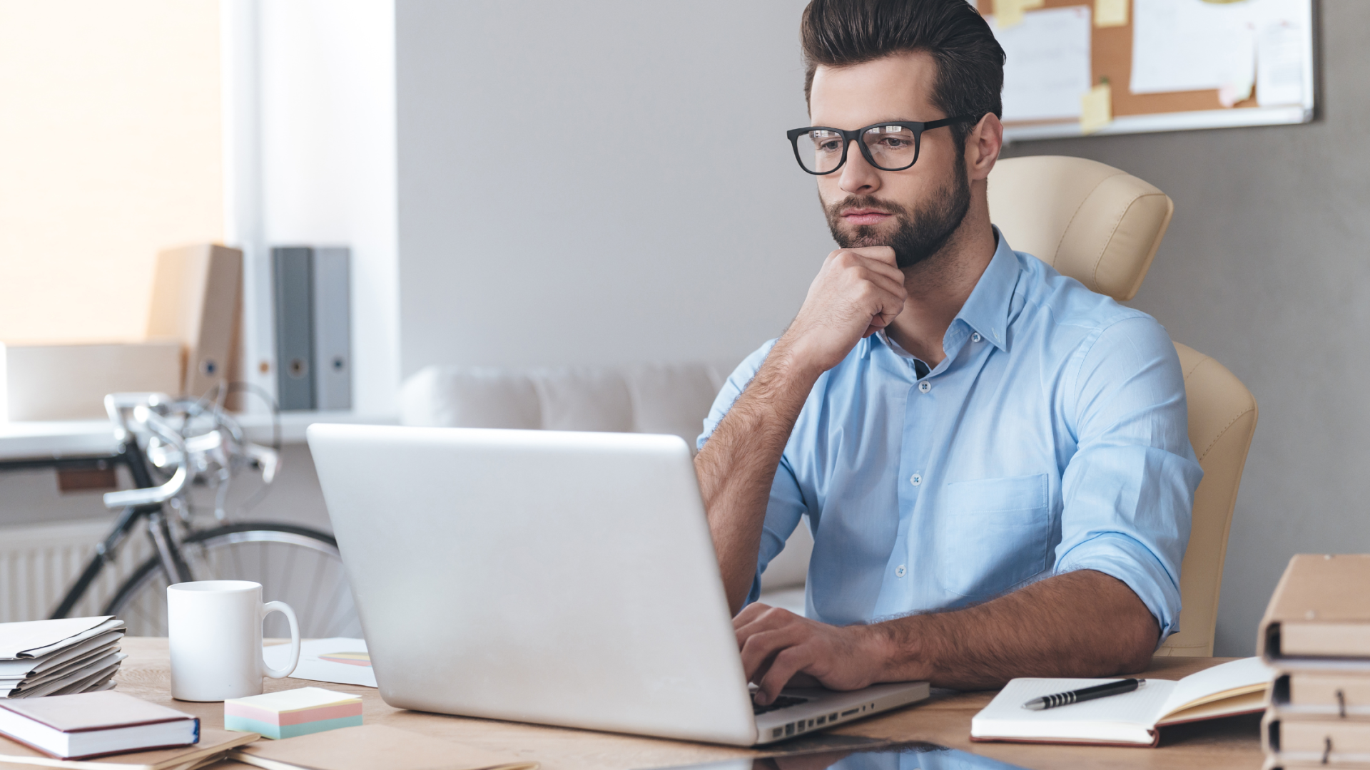 Man in a light blue shirt and glasses sits at a desk, thinking with his hand on his chin in front of an open laptop.